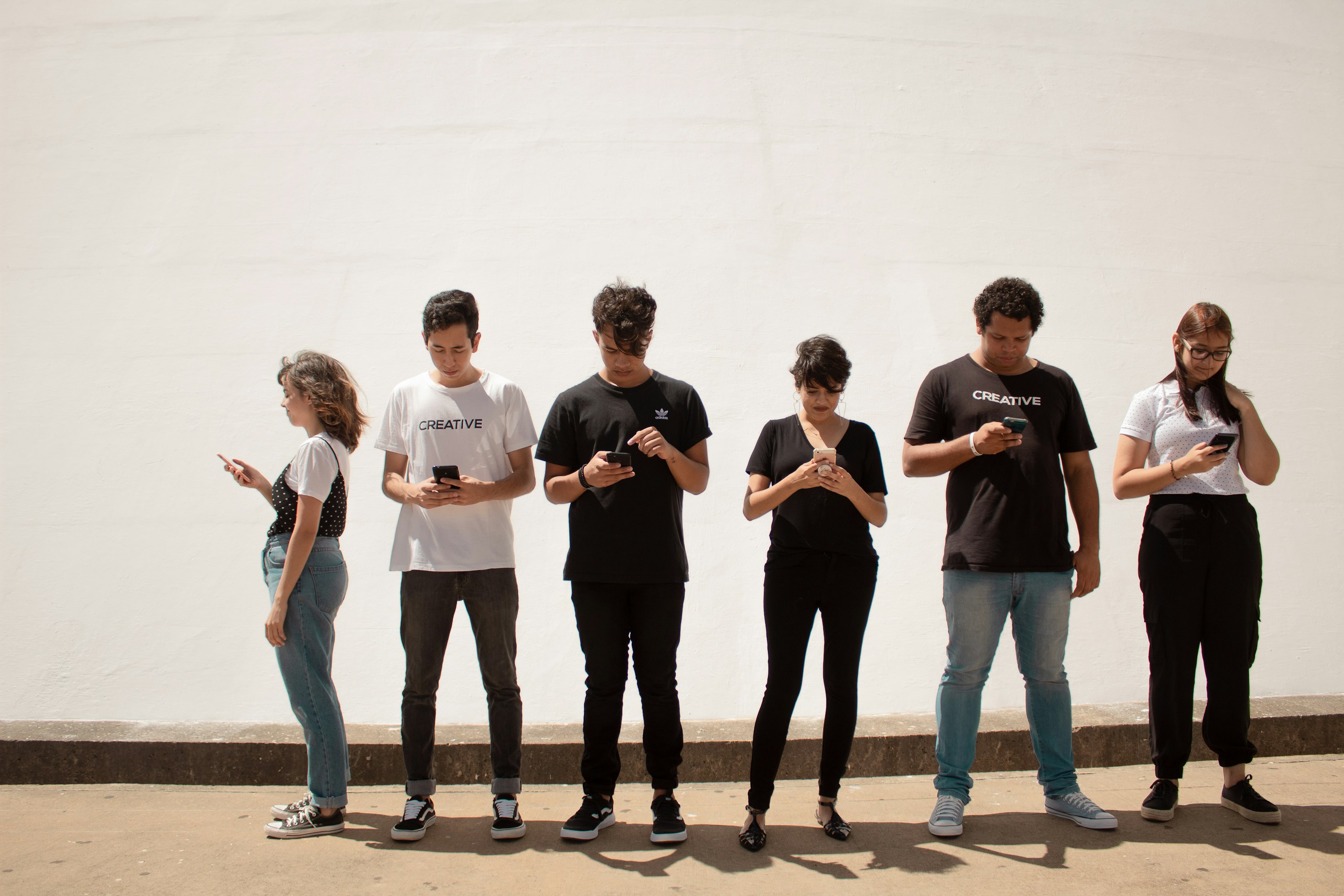 Six young people standing along a sunlit wall, each looking at their phone