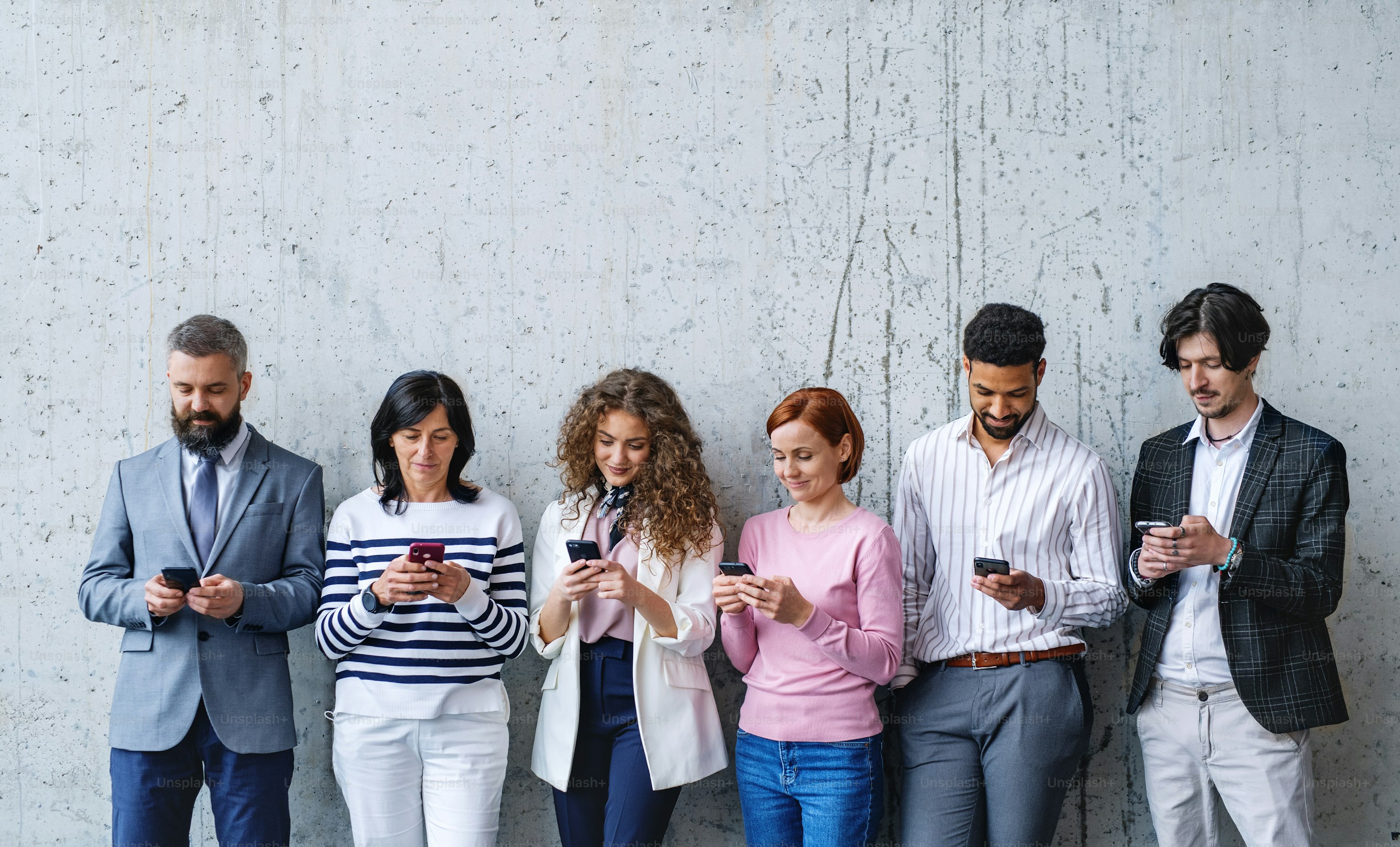 Six professionals in business dress standing against a concrete wall, each looking at their phone
